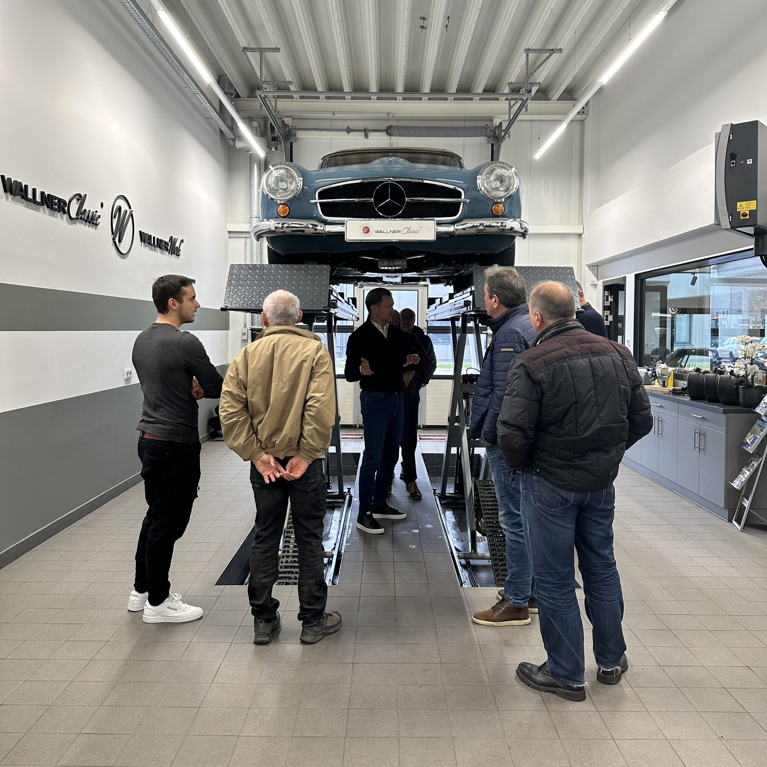 Zu sehen ist Herr Genz unter der Diagnosebühne. Ein blauer 190 SL befindet sich auf der Bühne. Weitere Personen begutachten das Fahrzeug.