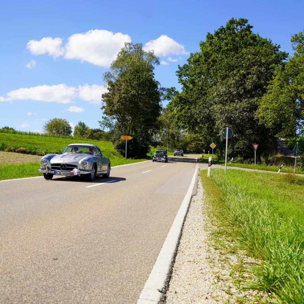 Zu sehen sind drei Oldtimer auf einer Landstraße. Grüne Wiesen und blauer Himmel.