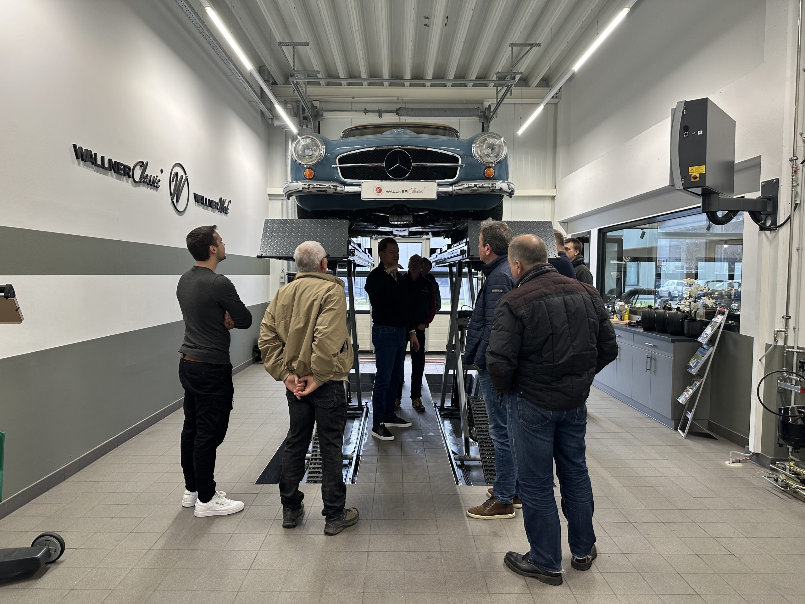 Auf dem Foto sind mehrere Personen die sich um einen blauen 190 SL aufstellen. Das Fahrzeug befindet sich auf einer Werkstatt-Hebebühne.