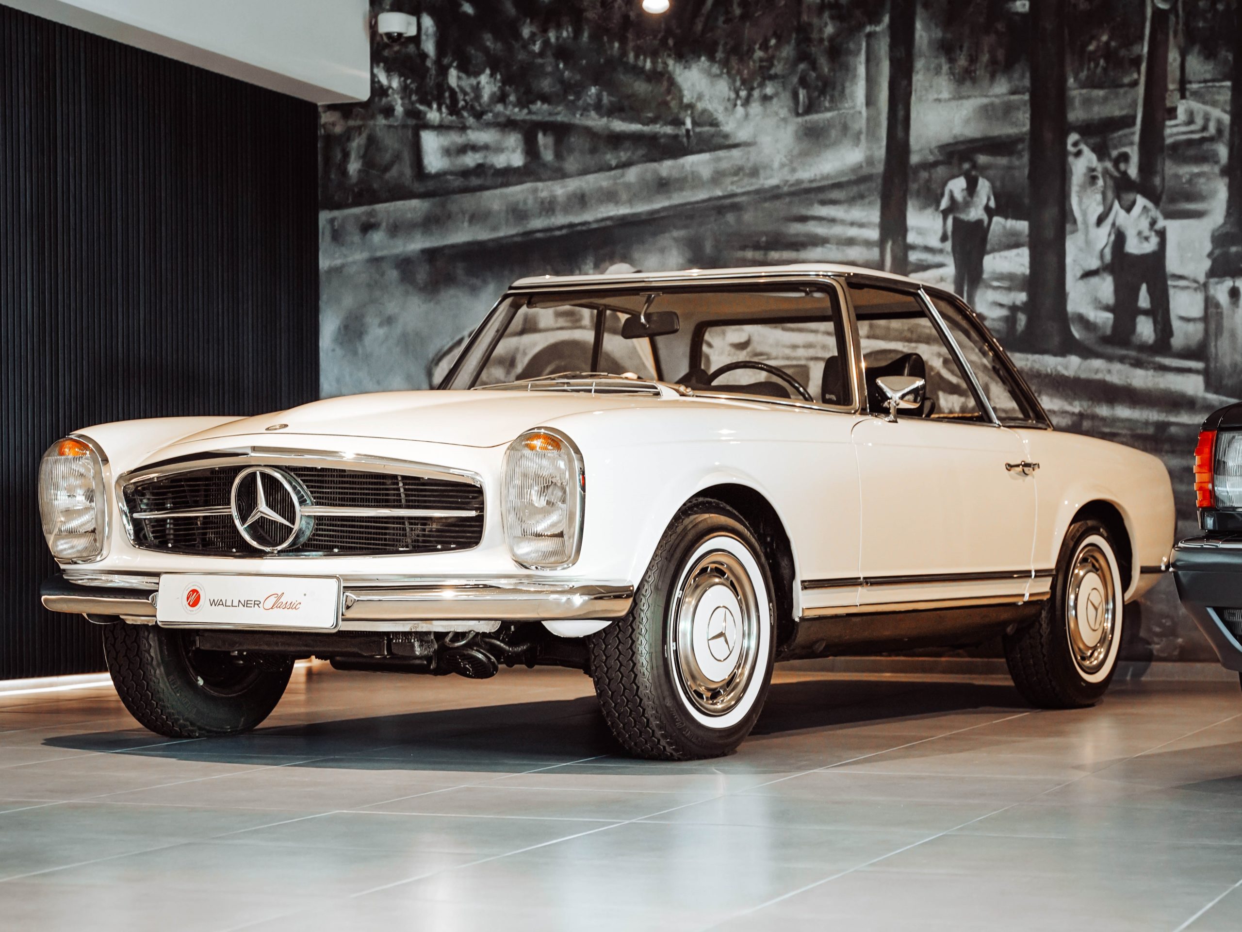 Zu sehen ist die Frontansicht einer weißen Mercedes Benz Pagode. Aufgenommen vor einer Wandmalerei in schwarz weiß. Im Ausstellungsraum von WALLNER Classic
