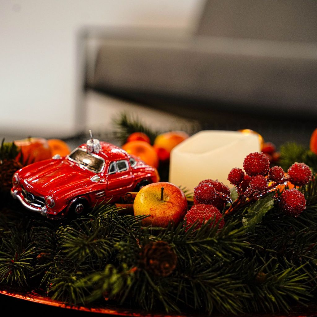 Zus sehen ist ein Adventskranz mit &Auml;pfeln, Beeren, einer wei&szlig;en Kerze, sowie einer roten Christbaumkugel in Form eines Mercedes 190 SL